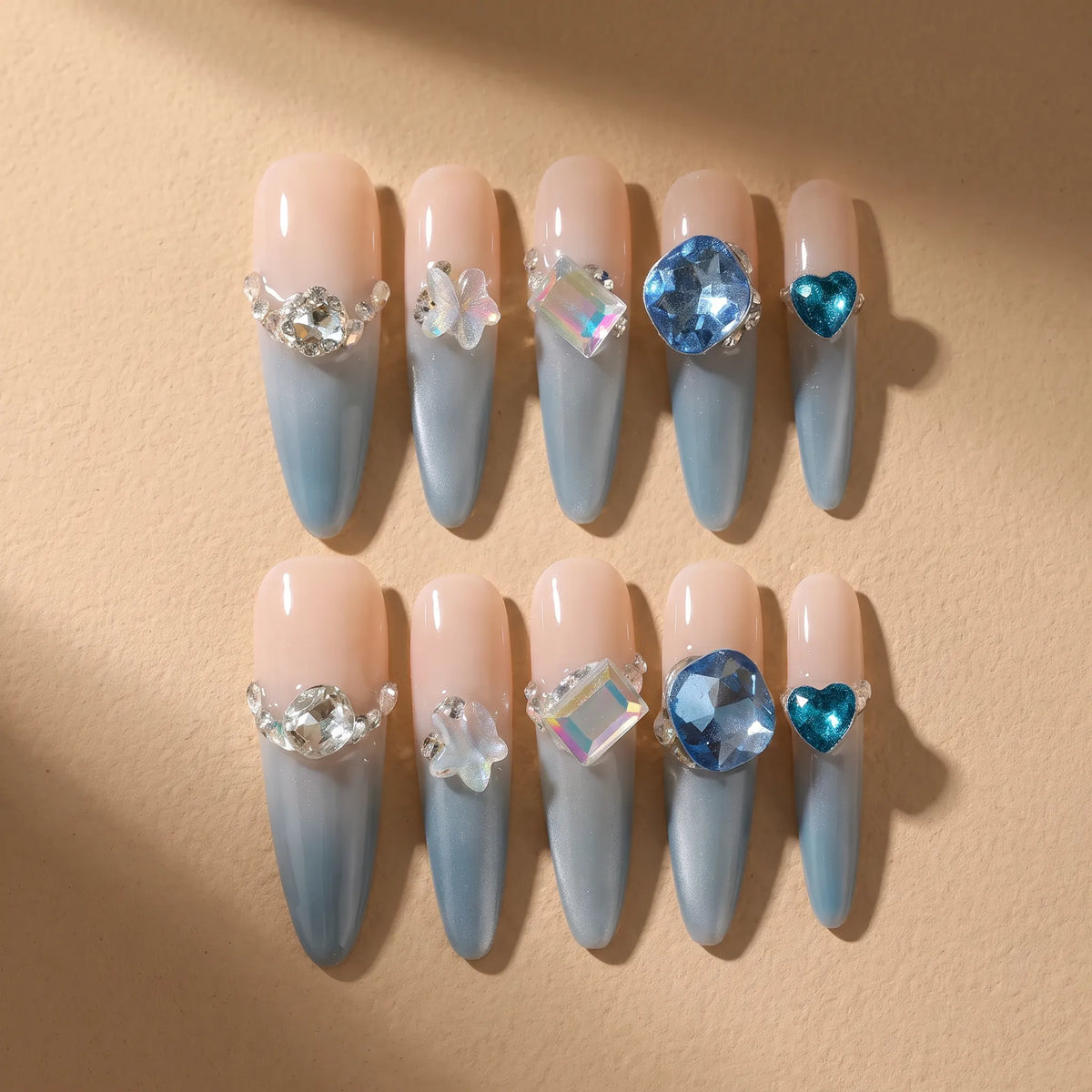 A set of 10 false press-on nails arranged in two rows on a beige background. The nails feature a gradient design with a pale peach-pink top half and a light blue-gray bottom half, decorated with various clear and blue rhinestones.